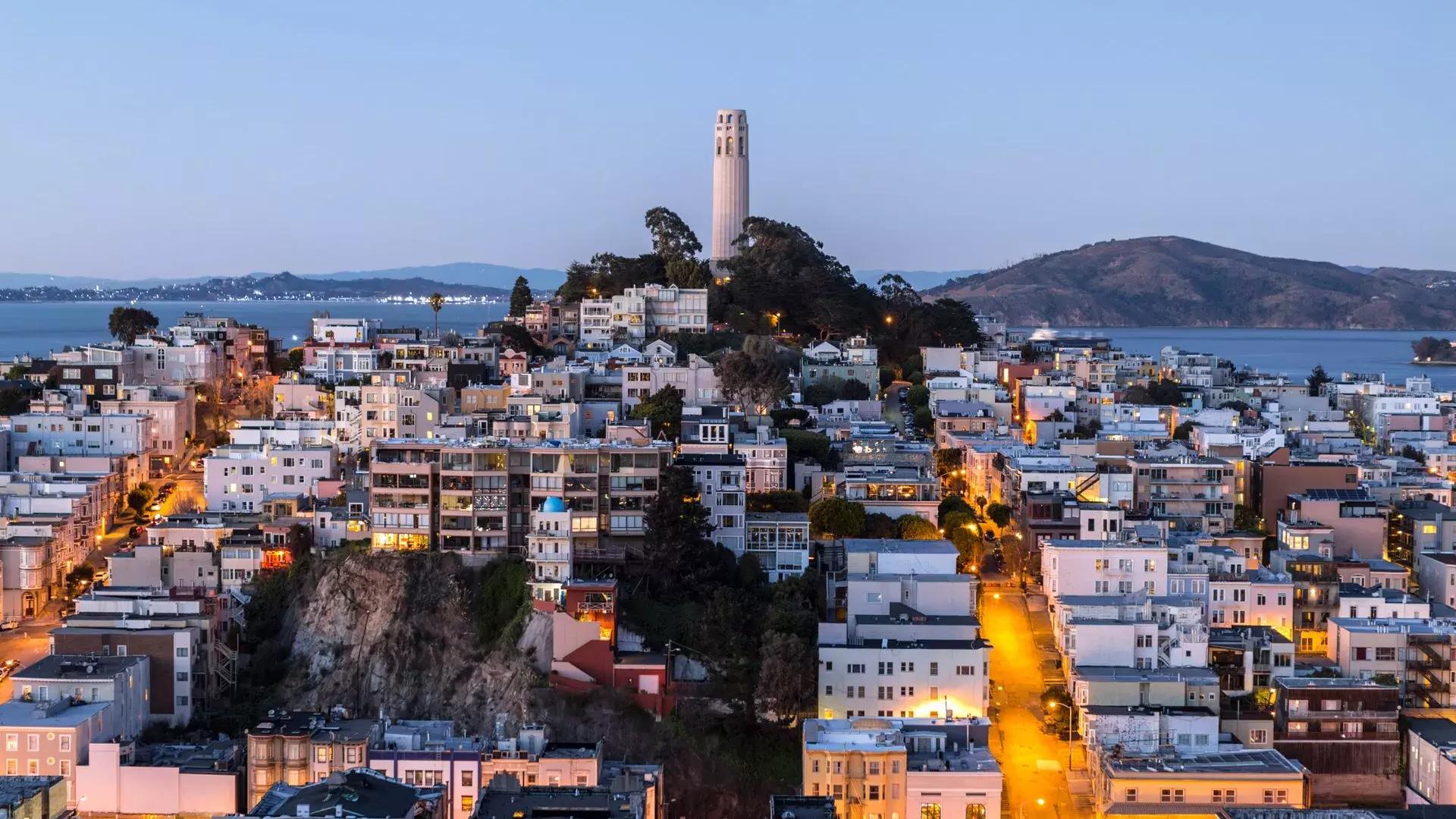 La Coit Tower de 贝博体彩app au crépuscule, avec des rues éclairées devant elle et la baie de 贝博体彩app derrière elle.